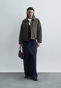 Brązowa, pudełkowa kurtka z złotymi guzikami i dwoma przednimi kieszeniami, noszona na granatowym stroju. Modelka trzyma ciemnoczerwoną torebkę.