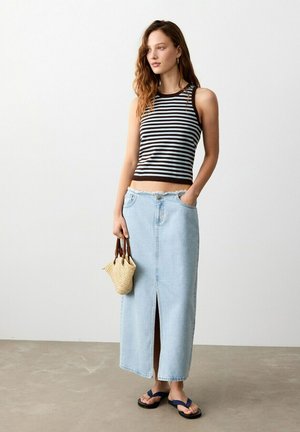 Jeune femme portant un haut sans manches à rayures noires et blanches et une jupe en jean bleu clair fendue à l'avant, tenant un sac tissé et des tongs.