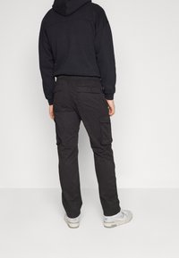 Person som bär svarta cargo-byxor, svart huvtröja och vita sneakers, stående vänd bort från kameran mot en enkel ljus bakgrund.