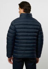 Mees, kes on pildil seljaga, kannab tumesinist vattimist jope tõstetud kraega ja tumedaid teksapükse ühevärvilise tausta ees.