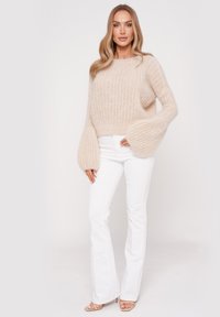 Beige oversized knit sweater with ribbed texture and bell sleeves, paired with fitted white flared jeans and light-colored sandals.