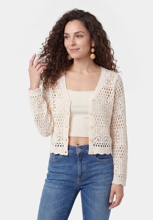 Donna con capelli ricci che indossa orecchini rotondi intrecciati, un cardigan aperto crema all'uncinetto sopra un crop top crema e jeans blu, posando con la mano vicino ai capelli.