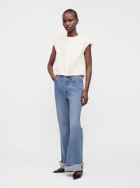 Femme debout portant un chemisier blanc sans manches, un jean bleu à jambes larges avec des ourlets effilochés et des talons noirs pointus sur un fond blanc uni.