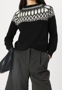 Svart tröja med ett vitt geometriskt mönster på oket, tillverkad av mjukt material, tillsammans med grå veckade byxor och en svart väska.