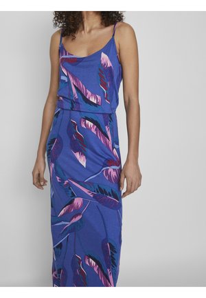 Kobieta w niebieskiej sukience bez rękawów z fioletowo-różowymi wzorami piór, stojąca na jednolitym jasnoszarym tle.