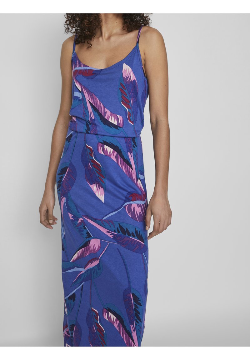 Femme portant une robe bleue sans manches avec des motifs de plumes violettes et roses, debout devant un fond gris clair uni.