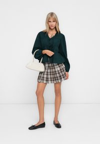 Kvinne som står i svart bluse, rutete skjørt, svarte leiligheter, og holder en hvit håndveske mot hvit bakgrunn.