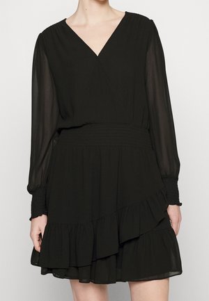 Czarna sukienka z długim rękawem, dekoltem w serek, marszczoną talią i mankietami, przezroczystymi rękawami oraz warstwowym falbanowym dołem, noszona przez stojącą osobę.