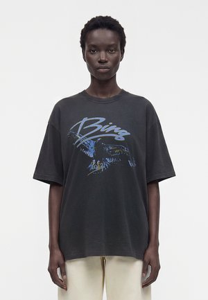 Personne debout aux cheveux courts portant un t-shirt noir oversize avec un motif d'aigle bleu et un pantalon beige sur un fond clair uni.