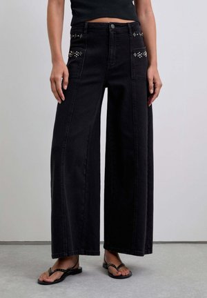 Pantalonii din denim negri cu picioare largi au aplicații decorative pe buzunarele din față, o talie înaltă și o textură solidă. Sunt asortați cu flip-flops.