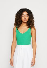 Bodysuit verde acanalado con un escote en V profundo, combinado con pantalones blancos de tiro alto. El bodysuit cuenta con tirantes anchos y una tela texturizada.