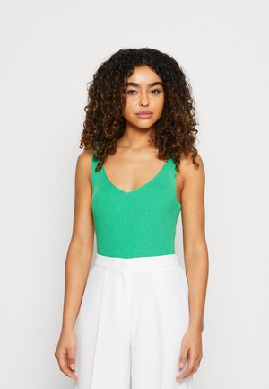 Green ribbed bodysuit with a deep V-neck, paired with high-waisted white trousers. The bodysuit features broad straps and a textured fabric.