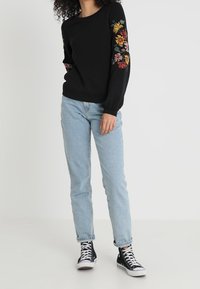 Black sweater with floral embroidery on sleeves, paired with light blue jeans and black high-top sneakers. Simple, casual design.