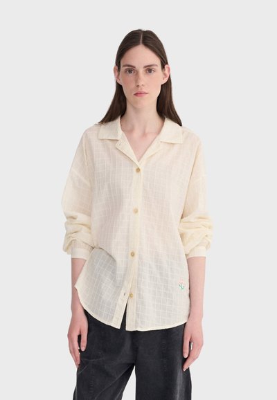 Femme aux cheveux bruns lisses portant une chemise ample couleur crème, boutonnée, avec les manches retroussées, et un pantalon noir, debout devant un fond blanc.