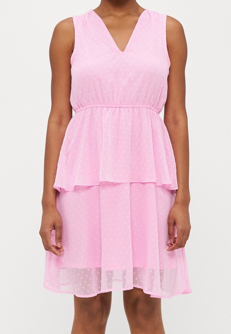 Robe rose sans manches avec col en V et jupe à volants, dotée d'un motif subtil à pois et d'une taille cintrée, portée par une personne debout devant un fond uni.