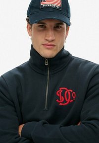 Sudadera de forro polar azul marino con cremallera y cuello alto, que presenta un logo rojo en el pecho y puños acanalados. Combinada con una gorra azul marino que muestra una bandera estadounidense.