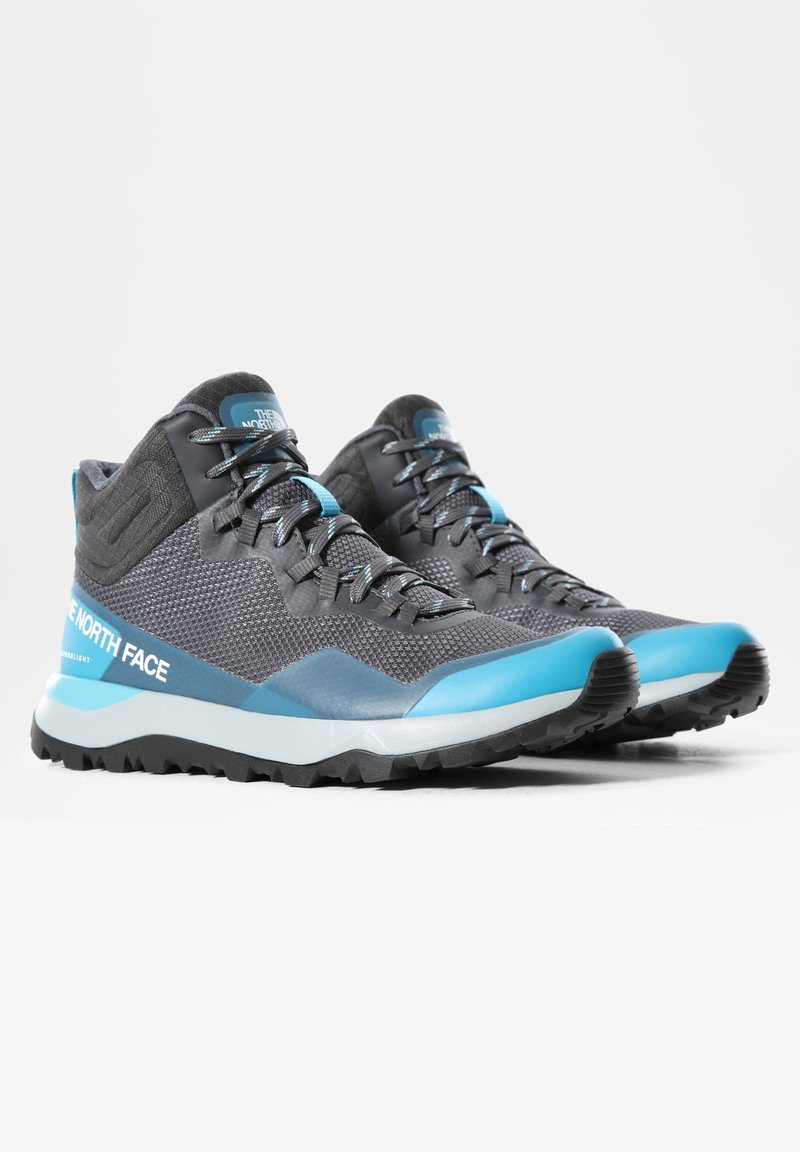 Paire de chaussures de randonnée montantes grises et bleues avec des lacets noirs et des semelles noires robustes, marquées "The North Face" sur le côté et la languette.