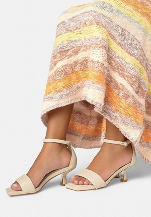 Pies con sandalias beige de tacón bajo y tiras en el tobillo, combinadas con una falda texturizada multicolor que presenta franjas horizontales en naranja, amarillo y rosa.