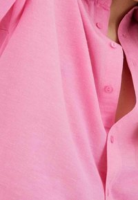 Camisa de botón rosa hecha de una tela texturizada, con un cuello y varios botones a juego a lo largo de la botonera.