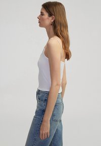 Witte katoenen tanktop met dunne bandjes, gecombineerd met lichtblauwe high-waisted jeans, weergegeven in een zijprofiel tegen een neutrale achtergrond.