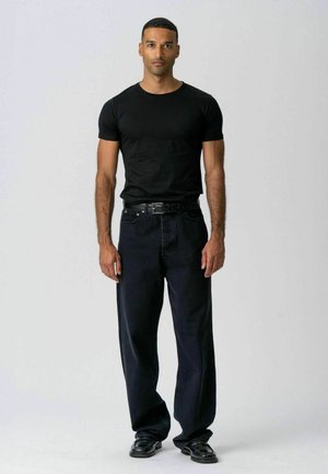 Mand stående iført en sort tætsiddende kortærmet skjorte, løse sorte bukser med bælte og sorte sko på en ensfarvet lys baggrund.