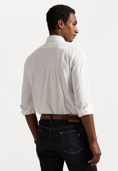 Λευκό πουκάμισο με μακριά μανίκια και διπλωμένα μανίκια, σε συνδυασμό με σκούρες μπλε τζιν παντελόνες, που διαθέτουν καφέ ζώνη και χαρακτηριστική ραφή στις πίσω τσέπες.