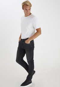 Schwarze Slim-Fit-Jeans mit einem dezenten Verblassen, kombiniert mit einem schlichten weißen T-Shirt. Das Model trägt schwarze Sneaker und posiert mit den Händen in den Taschen.