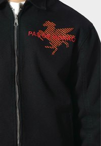 Schwarze Jacke mit einem silbernen Reißverschluss, mit einem orangefarbenen gestickten Design eines Pferdes und dem Text "PAS DE MONACO" auf der Brust. Strukturierter Stoff.