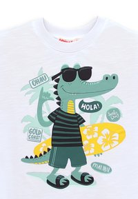 Tegneseriealligator med solbriller, stribet skjorte, shorts og sandaler, der står med et gult blomster-surfbræt og palmer i baggrunden.