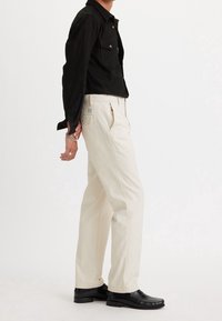 Svart skjorta med knappar och två framfickor, kombinerad med gräddfärgade byxor och svarta läderskor. Outfiten har en avslappnad passform.