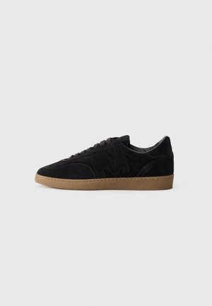 Schwarze Wildledersneaker mit strukturierter Oberfläche, einem markanten gebogenen Logo, grauen Schnürsenkeln und einer hellbraunen Gummisohle.