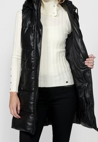 Femme portant un pull côtelé blanc et un pantalon noir tient ouvert un long manteau sans manches noir matelassé.