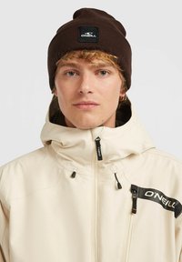 Cream-colored waterproof jacket with a high collar, black accents, and a side logo patch. Features a zip closure and adjustable hood. Brown knit beanie.