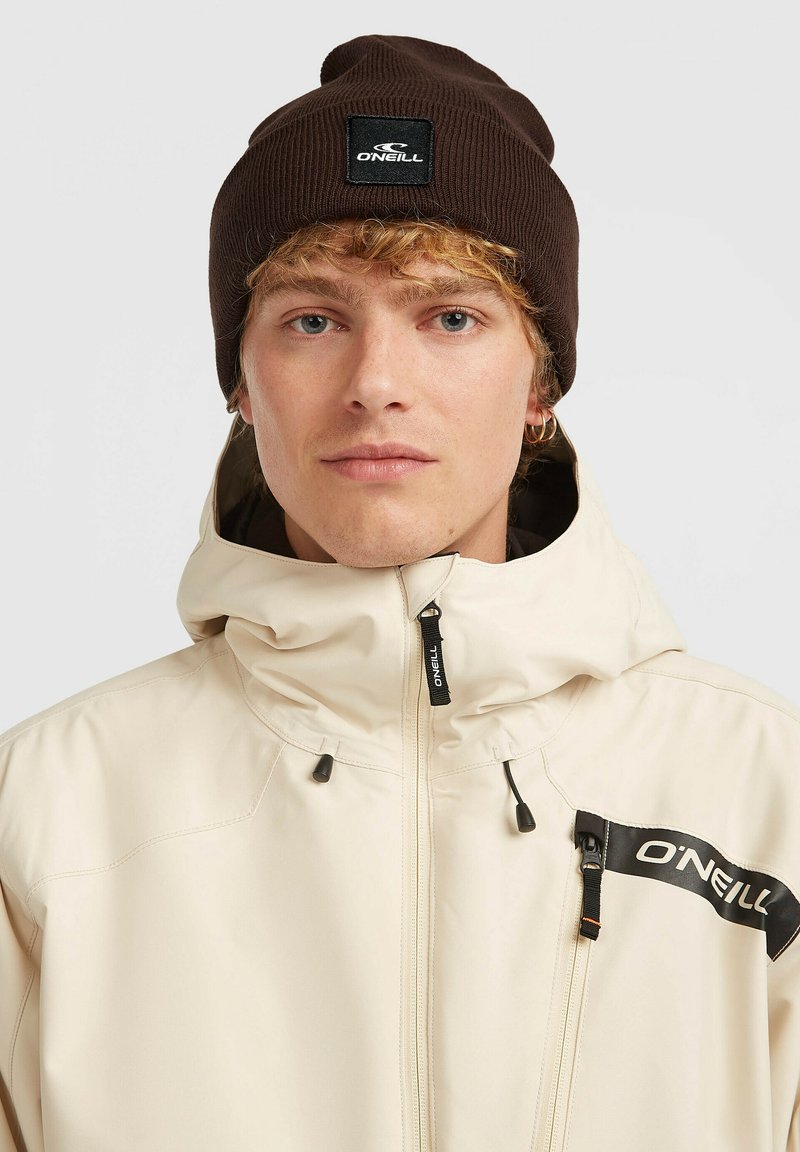 Beige zip-up jack met hoge kraag, met een zwart logo patch en accenten. Donkerbruine geribbelde beanie op het hoofd.