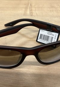 Aurinkolasit, joissa on ruskea muovikehys, tummennetut linssit ja tyylikäs muotoilu. Mukana hintalappu, jossa on viivakoodi ja UV-suojatiedot.