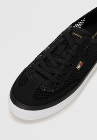 Baskets en mesh noir avec lacets noirs, semelle blanche et petit logo rectangulaire sur le côté sur fond blanc.