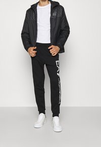 Černá větrovka s texturovanými rameny, bílý triko, černé tepláky se znakem na straně a bílé sportovní boty. Minimalistický design.