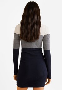 Femme aux longs cheveux bruns portant une robe moulante côtelée avec des bandes horizontales de couleur blanche, grise et bleu marine, debout de dos.