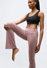 Γυναίκα που φοράει μαύρο αθλητικό μπλουζάκι και μωβ κολάν, τεντώνεται κρατώντας το ανυψωμένο γόνατό της με το ένα χέρι μπροστά σε λευκό φόντο.