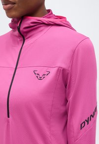 Άτομο που φοράει ροζ αθλητικό μπουφάν με μαύρο ημιδιαφραγμένο φερμουάρ, κουκούλα, μαύρο λογότυπο στο στήθος και κείμενο "DYNA" στο μανίκι.