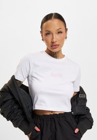 Biele krátke tričko s jemným ružovým logom, skombinované s čiernou bomber bundou. Model má na sebe čierne nohavice so sťahovaním a viditeľné tetovania.