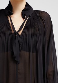 Blusa nera trasparente con texture plissettata, colletto smerlato e chiusura a fiocco. Il tessuto leggero crea una silhouette sfumata.