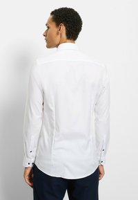 Weißes Langarmhemd mit figurbetontem Design, dezenter Textur und kontrastierenden dunklen Knöpfen an den Manschetten. Glatter Stoff mit abgerundetem Saum.