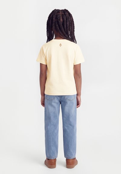 Παιδί με μακριά πλεξούδα που φοράει ανοιχτό κίτρινο κοντομάνικο πουκάμισο, μπλε τζιν και καφέ παπούτσια, στέκεται απέναντι, με την πλάτη, σε λευκό φόντο.