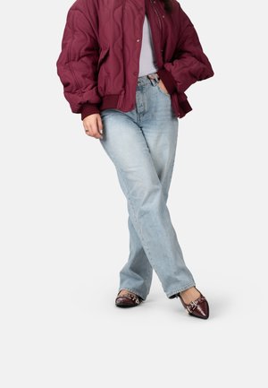 Person trägt eine bordeauxrote gesteppte Jacke, hellblaue Jeans und spitze bordeauxrote Schuhe mit metallischen Nieten und steht mit gekreuzten Beinen.