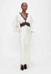 Gara balta satīna kleita ar melniem mežģīņu akcentiem, dziļo V veida izgriezumu un pūderveida piedurknēm. Kopā ar melniem augstpapēžu apaviem ar smailām galām. Gluda tekstūra.