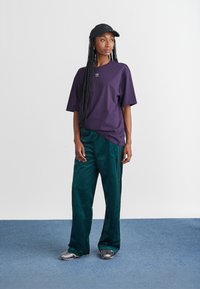 Purple oversized t-shirt with logo, green corduroy pants, and silver slip-on shoes, paired with a black cap, standing on blue flooring.