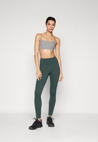 Grå sports-bh med tynne stropper, kombinert med høytaljede, mørkegrønne tights som har en jevn tekstur og en tettsittende fasong. Sorte joggesko.