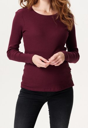 Femme portant un haut ajusté côtelé bordeaux à manches longues avec trois boutons sur le bas, associé à un pantalon noir, mains jointes.
