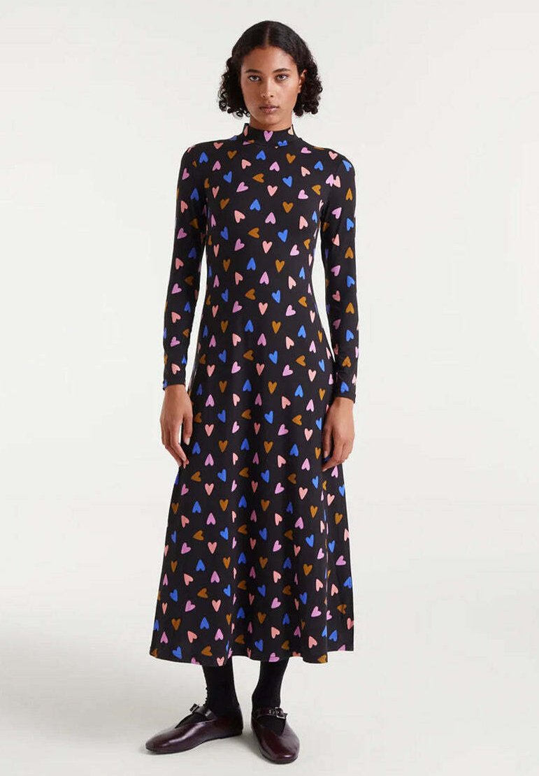 Mujer vestida con un vestido midi negro de manga larga con patrones de corazones coloridos, combinado con medias negras y zapatos marrón oscuro.